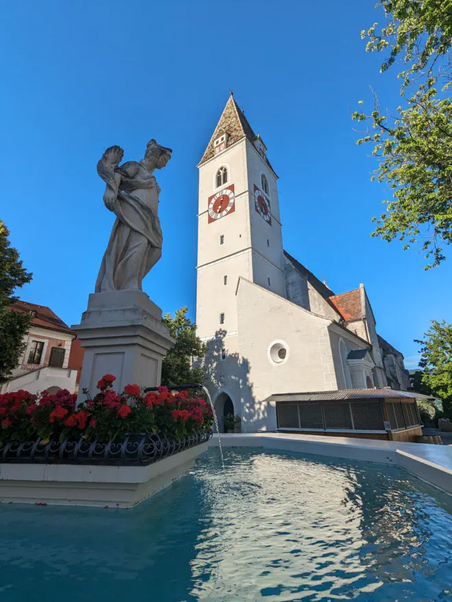 Brunnen am Spitzer Marktplatz und dahinter Kirchturm der Spitzer Kirche mit farbenprächtigem Dach